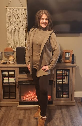 Reviewer in her living room wearing the Edna Hooded Cardigan over a matching taupe turtleneck and dark leggings.