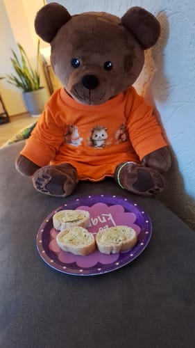 Recensent's Verzwaringsknuffel Beer in een oranje shirt, zittend op een bank met een bordje ervoor.