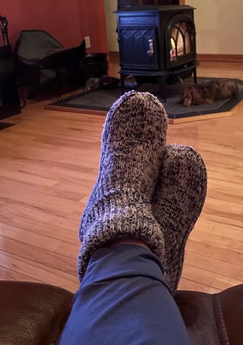Customer relaxing with feet up, wearing cozy speckled Gripper Slipper Booties in front of a warm fireplace.