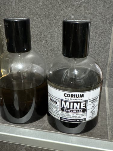 Il recensore mostra il suo profumo Corium accanto a un altro profumo su uno sfondo grigio.