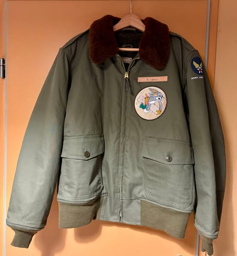 Reviewer's customized olive USAAF Type B-10 Flight Jacket hanging on a hanger against an orange wall.