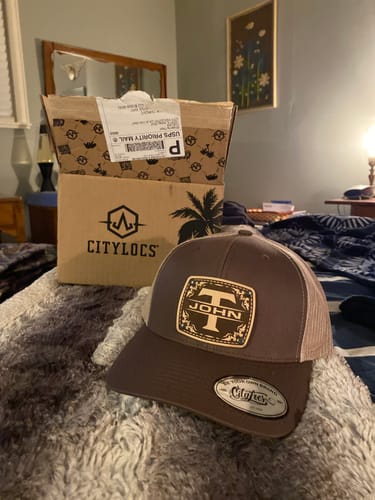 Customer's brown Monogram Custom Hat with a personalized leather patch showing the initial 'T' and the name 'JOHN', resting on a bed.