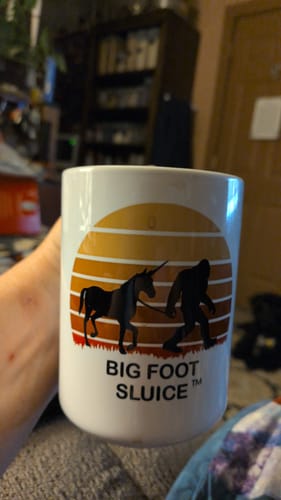 A customer holds the Bigfoot white glossy mug, which has a graphic of Bigfoot leading a unicorn.