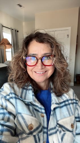Reviewer smiling while wearing her stylish, red Iris Blue Light Readers in her home.