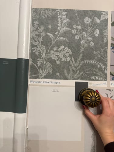 Customer planning a room with a sample of the floral Winsome Wallpaper, paint swatches, and a vintage-style brass doorknob.