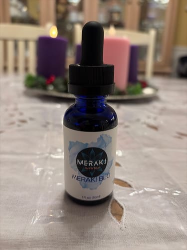 Customer's blue bottle of Meraki Blu with a black dropper on a table, with lit candles in the background.