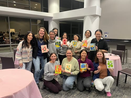 A group of happy reviewers at a painting event holding up their completed Mini Paintings of fruit.