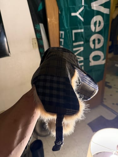 Customer's hand holding the plaid trapper hat, showing the faux fur-lined earflaps and strap.