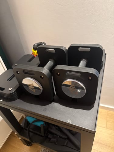 Kunde zeigt seine zwei schwarzen, verstellbaren QUAD Kurzhanteln auf einem Ablageregal.