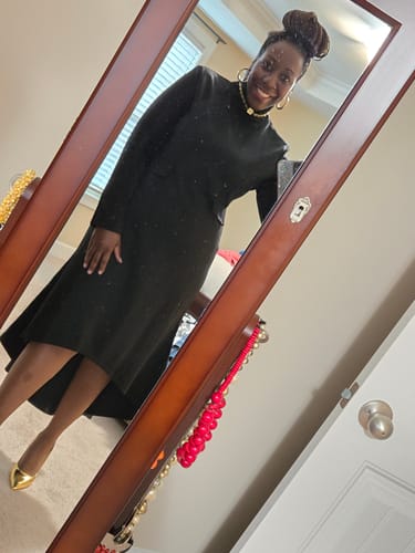 Reviewer smiling in a mirror wearing the black long-sleeved high neck midi dress, showing its fit and high-low hem.