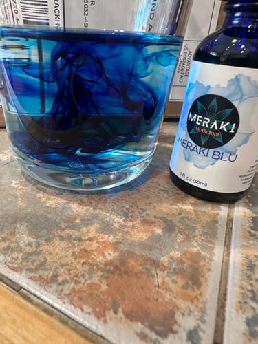 Reviewer's bottle of Meraki Blu next to a glass showing the dark blue dye mixing with water.