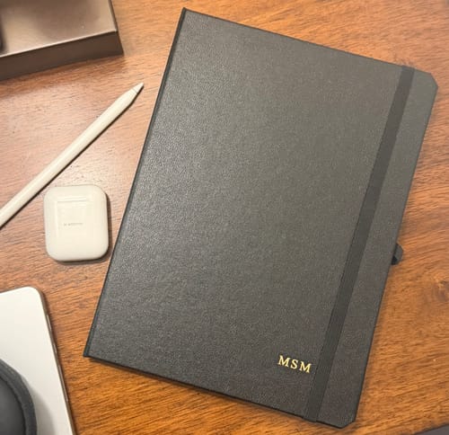 Customer's monogrammed Classic Black iPad Case shown closed on a wooden desk next to an Apple Pencil and AirPods case.