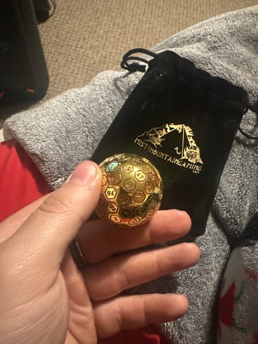 Reviewer holding the large, gold Orb D100 metal die in their hand, with the black velvet bag in the background.
