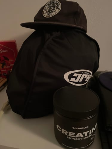 Customer's black tub of Complex Creapure Creatine on a white shelf, next to a sports bag and a cap.
