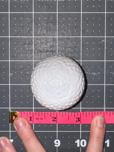 Customer measures the opaque golf ball with a pink tape measure on a gridded mat to show its large size.