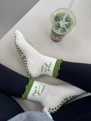 Reviewer wearing the white Pilates & Matcha socks with green ruffled trim and sole grips, seated with an iced matcha drink.