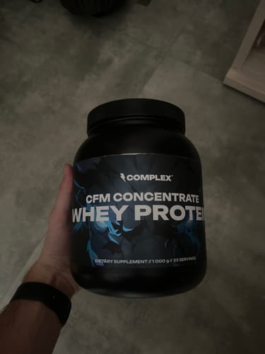 Customer holding a large black tub of Complex CFM Concentrate Whey Protein indoors.