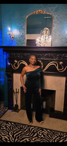 Customer posing in the dark green Nightfall One Shoulder Velvet Jumpsuit in front of a fireplace.