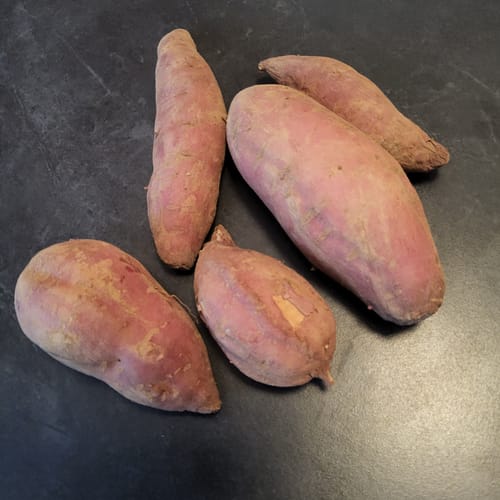 Rezensent zeigt fünf frische, violette 'Okinawa' Süßkartoffeln in unterschiedlichen Größen auf einem dunklen Tisch.