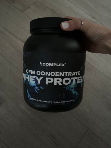 Customer's hand holding a large black tub of Complex CFM Whey Protein over a wooden floor.