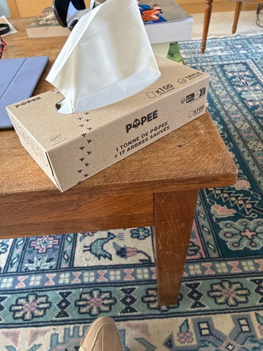 Un client montre la boîte de mouchoirs en carton posée sur une table en bois.
