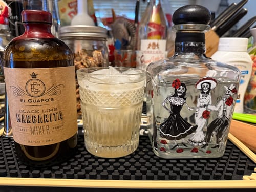 Customer's bottle of Black Lime Margarita Mixer next to a prepared margarita in a glass and a bottle of tequila on a bar.