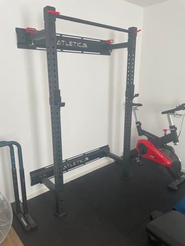 Rezensent zeigt das schwarze ATLETICA Wall Rack R7, das in einem Home-Gym an einer weißen Wand montiert ist.