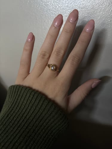 A customer's hand wearing the delicate Vintage Royal Pearl Ring on their ring finger, with a green sweater cuff visible.
