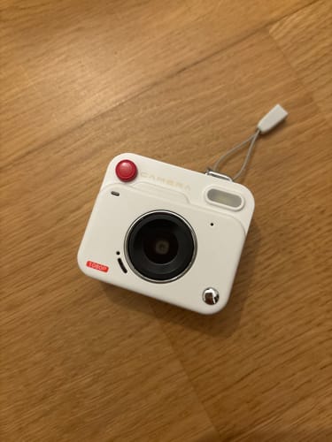 A reviewer's white RetroShot-Pro Mini camera with a red button, resting on a light-brown wooden surface.