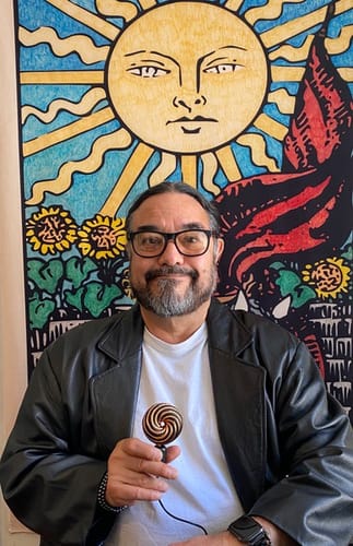 Reviewer smiling while holding the spiral-patterned Qi Coil™ Mini in his hand in front of a colorful tapestry.