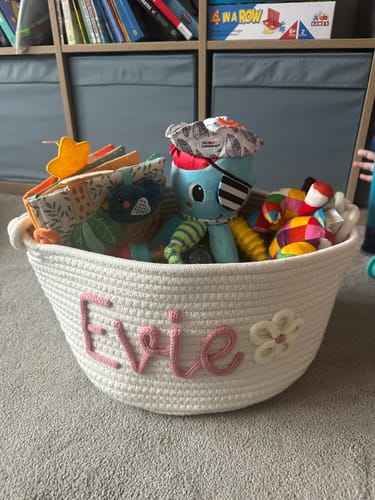 Customer's personalized knitted basket with 'Evie' in pink and a flower icon, filled with baby toys on a floor.