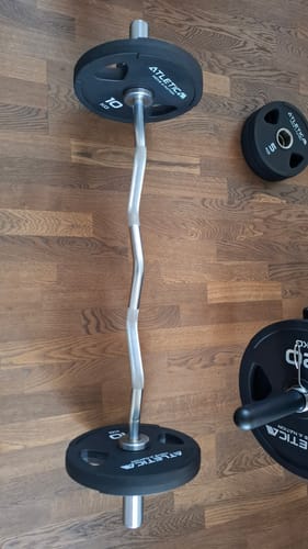 Rezensent zeigt die ergonomische SZ-Stange mit Gewichten auf einem Holzboden.