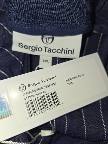 Reviewer's close-up of the Gessato track pant showing a size mismatch: the sewn-in tag is 3XL and the paper tag is 2XL.