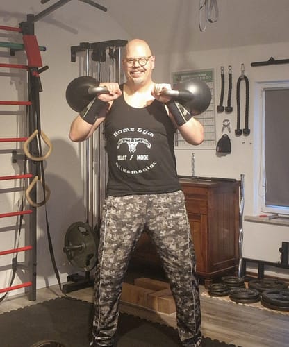 A smiling reviewer holds two Adjustable Kettlebells at shoulder height in a home gym.