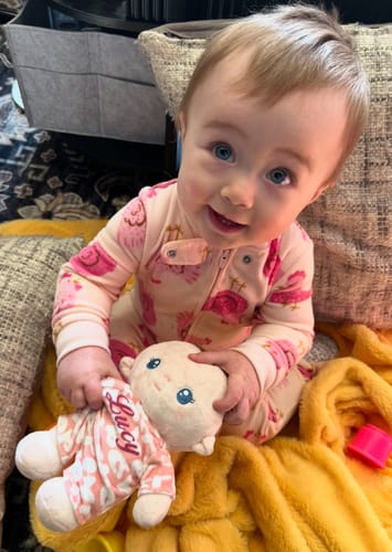 Customer's smiling baby holding their soft Personalized Pudding Baby Girl doll, which has 'Lucy' embroidered on its outfit.