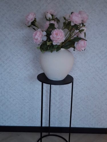 Recensent toont de roze zijden pioenrozen in een witte vaas op een hoge, zwarte standaard.
