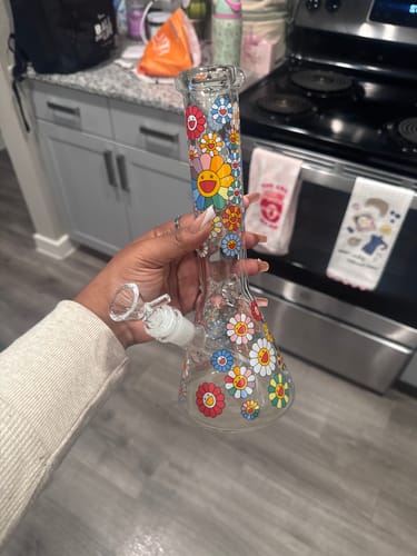 Reviewer holding the Happy Flowers Beaker Water Pipe, showing its colorful smiling flower decals in a kitchen.