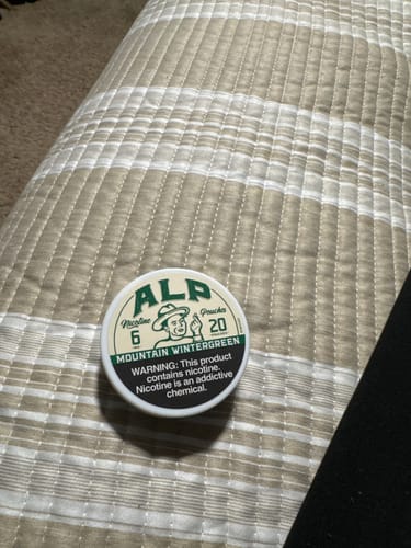 Customer's tin of ALP Mountain Wintergreen Nicotine Pouches resting on a striped surface.