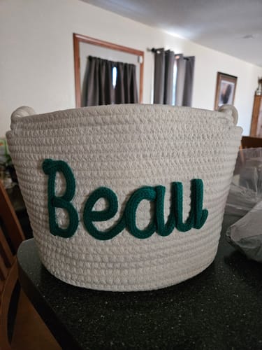 Customer's white knitted basket personalized with the name 'Beau' in dark green yarn.