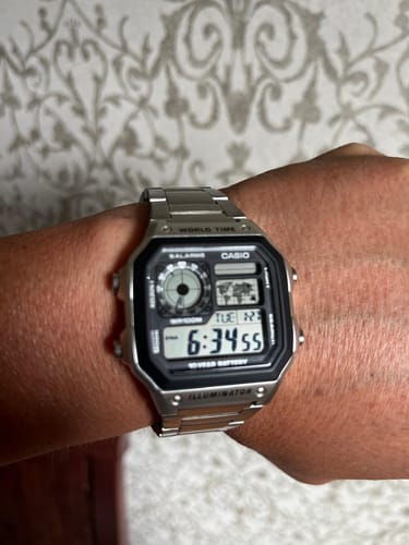 Cliente mostrando su reloj Casio World Time Royale plateado en la muñeca contra un fondo estampado.