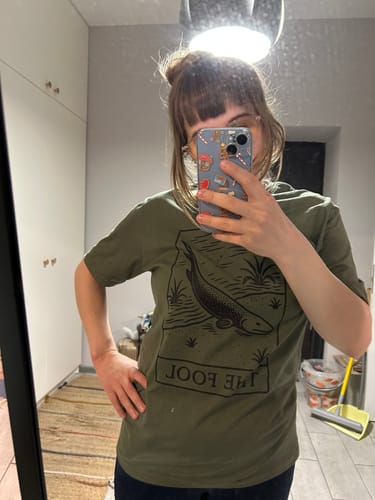 Reviewer taking a mirror selfie wearing the olive green Devonian Fool T-Shirt, showing the fit and graphic.