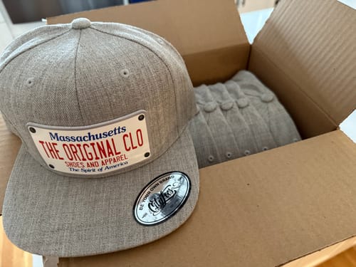 Customer's custom Massachusetts Plate Hat resting on an open box. The light grey hat features a patch for 'THE ORIGINAL CLO'.