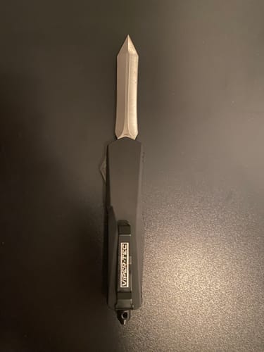 Reviewer's Black Ghost OTF knife with its sharp Spartan blade fully extended on a dark surface.