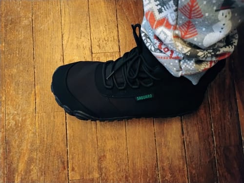 Reviewer wearing the black Will II - Winter Barefoot Shoes with festive pants on a wooden floor.