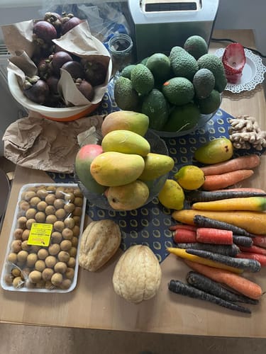 Rezensent zeigt eine große Auswahl frischer Früchte und Gemüse auf einem Tisch, Teil einer Bewertung für Rambutan.