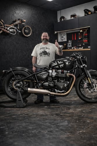 Client portant le t-shirt de la COLLECTION GENTLEMEN et posant à côté d'une moto dans un garage.