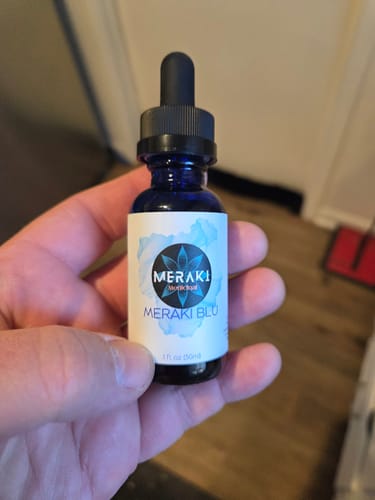 A customer holds the blue glass bottle of Meraki Blu, showing its label and dropper.