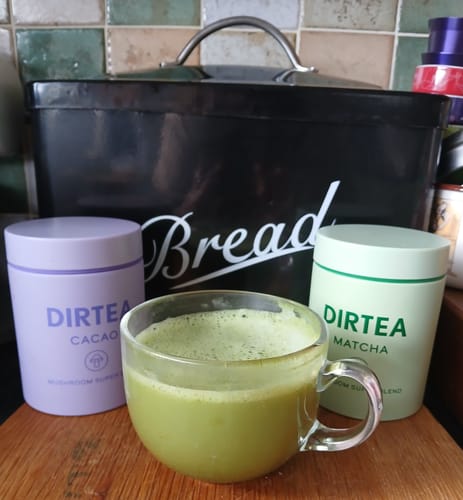 Reviewer's Rise + Unwind set with DIRTEA Cacao and Matcha canisters next to a prepared green matcha drink in a clear mug.