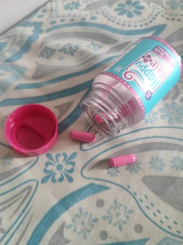 Cliente mostra o pote aberto de Happy Hair com algumas cápsulas rosas espalhadas sobre um tecido estampado.