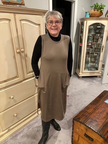 Reviewer wearing the sleeveless taupe mystery dress layered over a black turtleneck and tall boots in a living room.